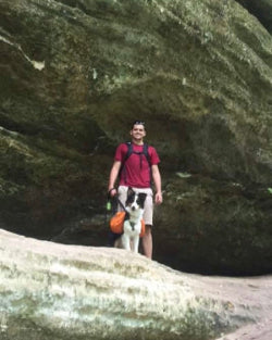 Man with a dog standing on a rock formation with a large rock in the background